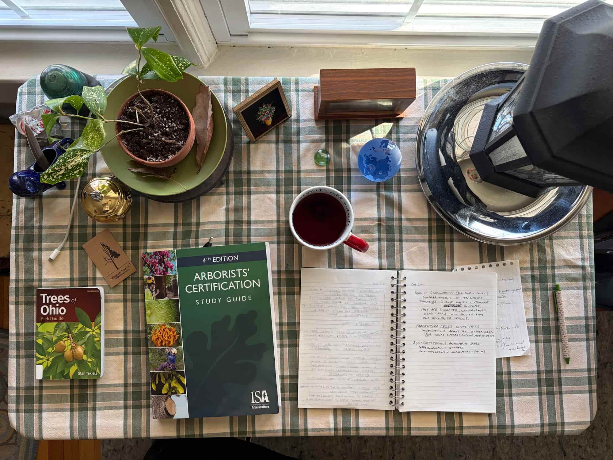 The ISA Arborists' Certification Study Guide open on a desk beside a notebook of handwritten notes, with ArbAssist on a laptop